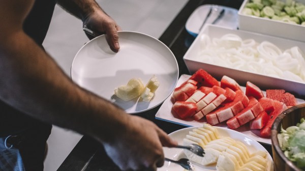 Person serving a plate with slices of melon and watermelon from a buffet of fresh fruits and vegetables.