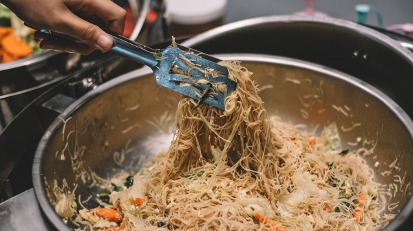 Hand holding tongs with cooked noodles above a pot, mixed with vegetables.