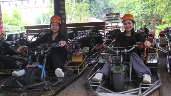 Two people in helmets sitting on go-karts in a shaded area with trees in the background.