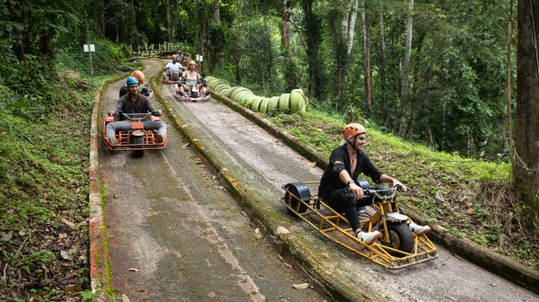 People riding luge carts down a forest path wearing helmets and protective gear.