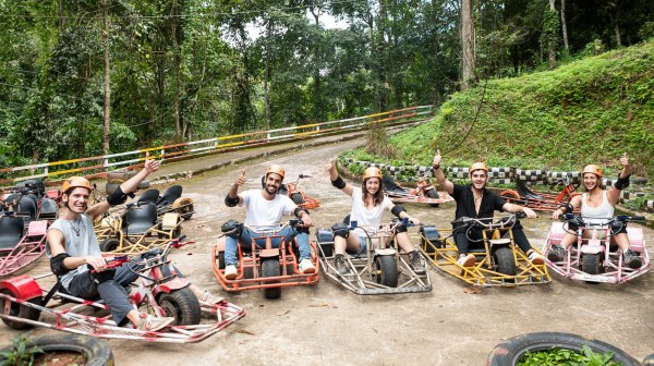 Group of six people in helmets sitting on go-karts, giving thumbs up, with forest background.