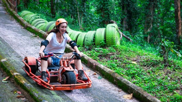 Woman riding a small red go-kart down a forest path with green tires lining the side.