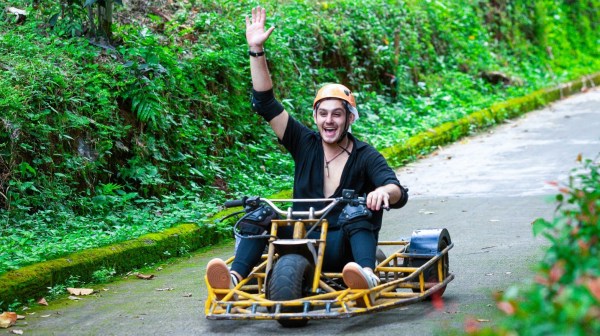 Person joyfully riding a go-kart on a green, leafy path, waving with one hand.