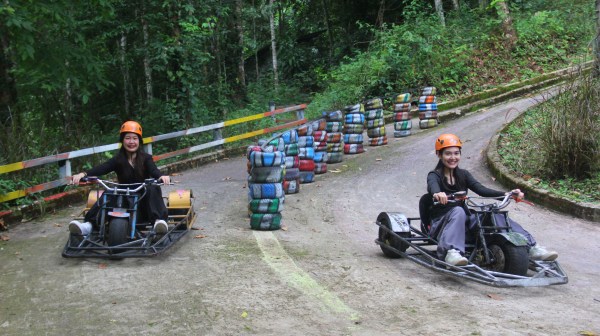 Two people in helmets riding go-karts down a wooded path with colorful tires as barriers.