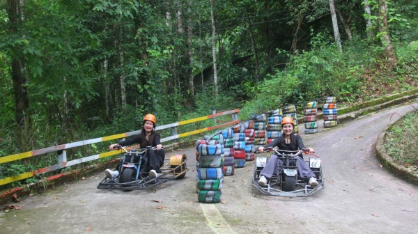 Two people in orange helmets ride go-karts on a forest track with colorful tires.