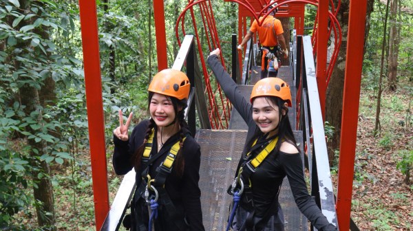 Two people with helmets on a metal bridge set in a forest, posing under red arch structures.