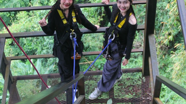 Two people in harnesses and helmets smile on a metal staircase amidst lush greenery.