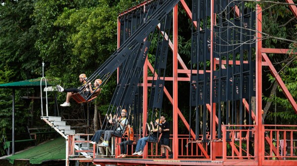 Four people on a red and black adventure swing structure in a forest setting.