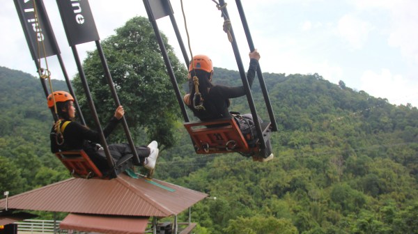 Two people with helmets on zipline swings over a lush green forest landscape.