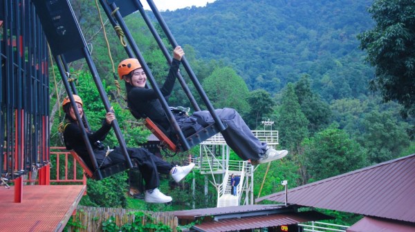 Two people on a swing ride wearing helmets, with a forested mountain backdrop.