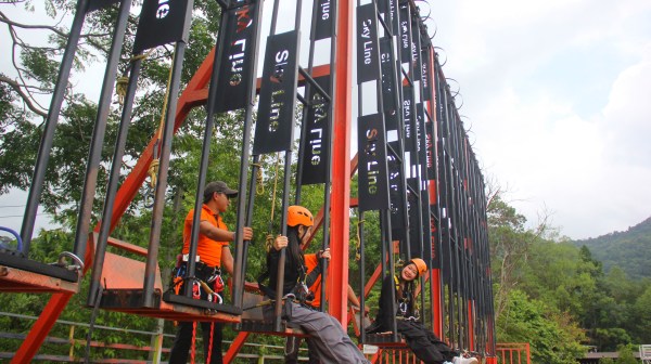 Three people on a swing ride, wearing helmets and harnesses, with trees and sky in the background.