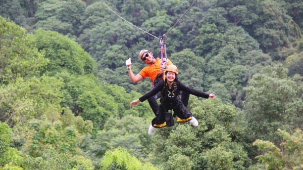 Two people ziplining over a lush green forest, smiling and wearing helmets.