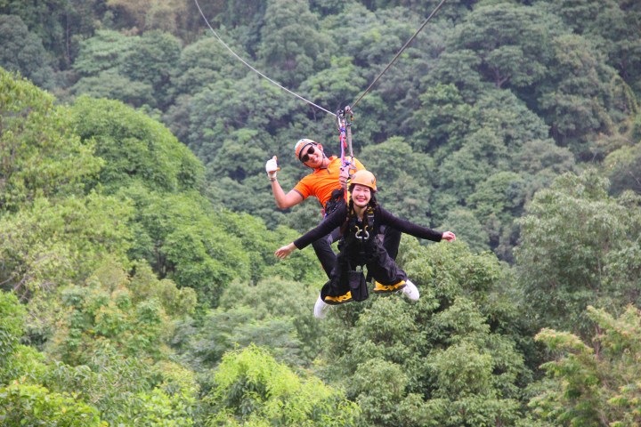 Two people ziplining over a lush green forest, smiling and wearing helmets.