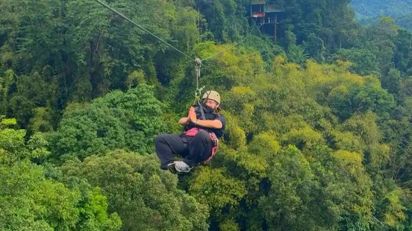 Person ziplining above lush green forest with cloudy sky background.