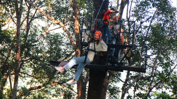 Person in orange helmet zip-lining in a forest with others on a platform.