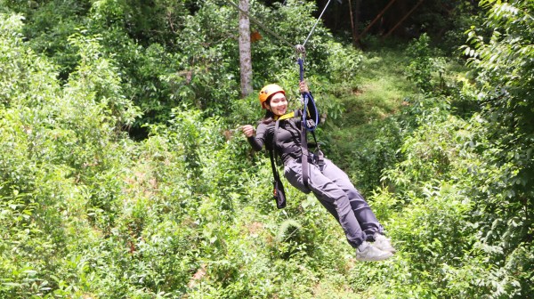 Person ziplining through a lush green forest wearing a helmet and safety gear.