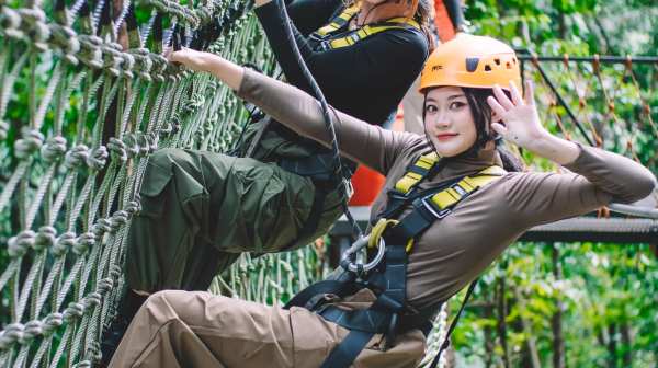 Two people in helmets smiling and climbing a rope net bridge in a forest.