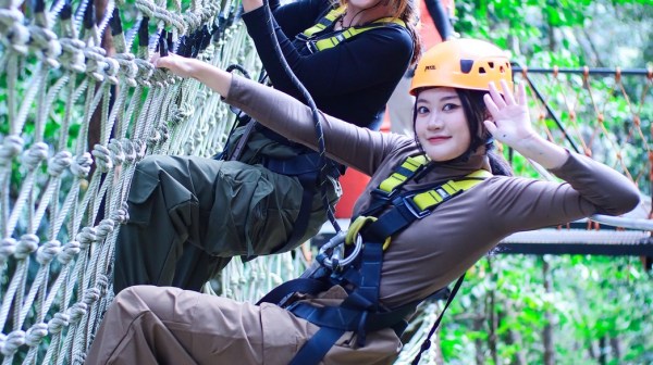 Two women in helmets climbing a rope net in a forest adventure park.