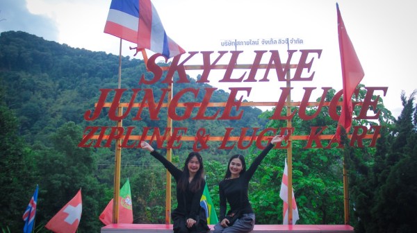 Two smiling people sitting under a sign reading 'SKYLINE JUNGLE LUGE' with flags and trees in the background.