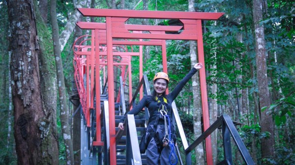 Person in helmet smiles on staircase with red archway, surrounded by forest trees.